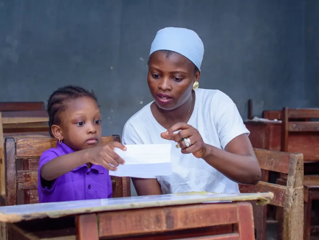 A young girl and a woman seated at wooden desks in a classroom, with the woman guiding the child through reading or writing on a piece of paper.