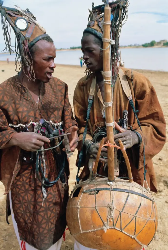 wo Malian griots in traditional attire performing by the riverside in Sofara, Mali, with one playing a stringed instrument made from a calabash.