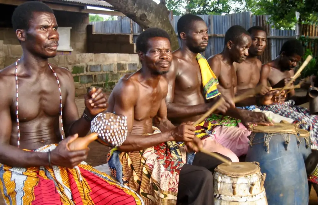 A group of African men in traditional clothing performing a drum ensemble outdoors, playing various percussion instruments including shekere and hand drums.